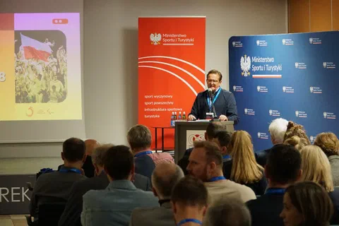 Mówca przy mównicy przed uczestnikami konferencji i ekranem ze zdjęciem reprezentacji.