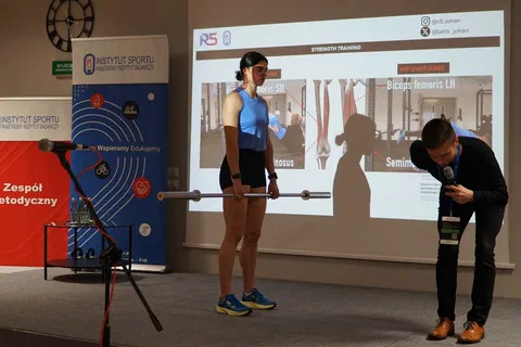 Pokaz ćwiczenia ze sztangą na scenie przed ekranem projekcyjnym i roll-upami Instytutu Sportu – PIB.