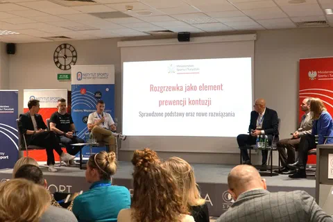 Czworo uczestników panelu dyskusyjnego siedzących na scenie po obu stronach ekranu z prezentacją o rozgrzewce.