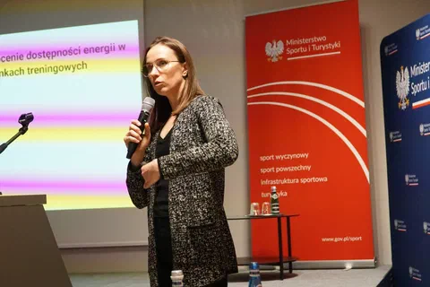 Kobieta w okularach wygłaszająca prelekcję o dostępności energii na tle banerów resortu.