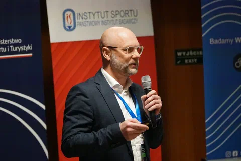 Prelegent w okularach przemawiający do mikrofonu na tle banerów Instytutu Sportu – PIB.