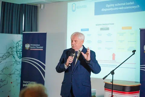 Prelegent w garniturze przemawiający do mikrofonu na tle ekranu ze schematem badań diagnostyczno-wdrożeniowych.