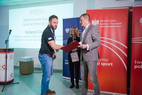 Wręczenie dyplomu w bordowej oprawie trenerowi na tle banerów Ministerstwa Sportu i Turystyki.