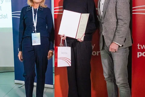 Trzy osoby pozujące z wręczonym dyplomem na tle czerwono-niebieskich banerów resortu.