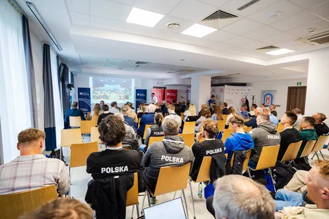 Widok z tyłu sali na uczestników konferencji w koszulkach z napisem „Polska”.