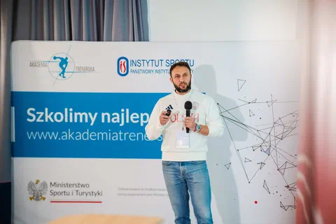 Prelegent z mikrofonem występujący na tle ścianki Akademii Trenerskiej i Instytutu Sportu – PIB.