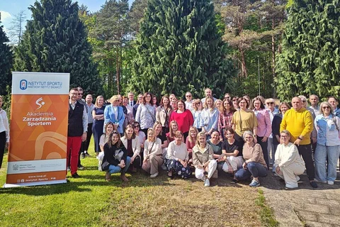 Grupowe zdjęcie uczestników szkolenia w plenerze na tle zieleni i baneru Akademii Zarządzania Sportem.