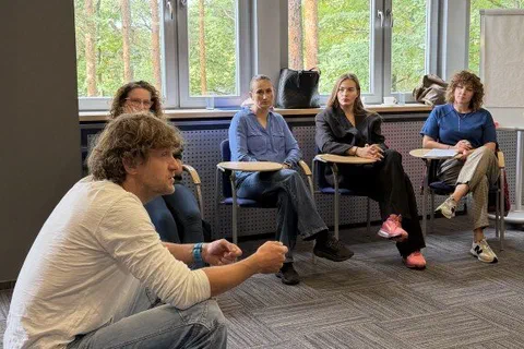 Prowadzący kucający obok trzech uczestniczek i rozmawiający z nimi podczas ćwiczenia grupowego.