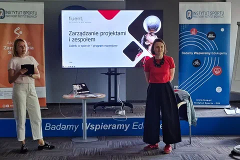 Dwie prowadzące stojące po obu stronach ekranu z tytułem szkolenia „Zarządzanie projektami i zespołem”.