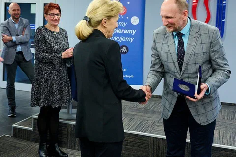Wręczenie medalu w otwartym etui i uścisk dłoni na tle ścianki z logo Instytutu Sportu