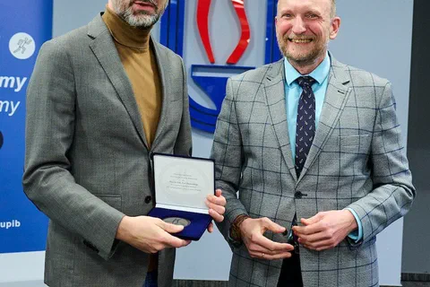 Dwaj mężczyźni w marynarkach pozujący z przyznanym medalem w etui przed logo Instytutu