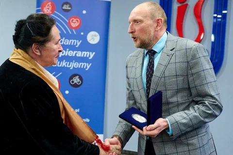 Wręczenie medalu Instytutu Sportu kobiecie w czarnej marynarce i szalu na tle roll-upu z hasłem Badamy Wspieramy Edukujemy
