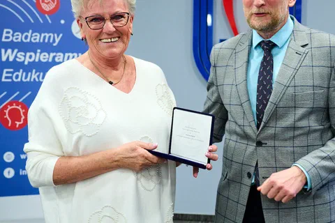 Kobieta w jasnej bluzce prezentująca medal w etui obok wręczającego podczas uroczystości