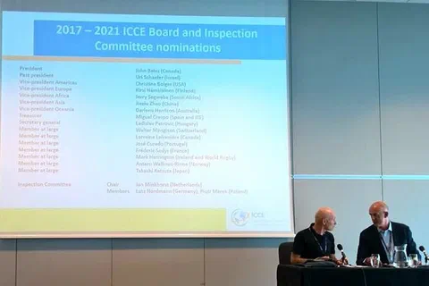Dwaj prowadzący przy stole prezydialnym pod ekranem z listą nominacji do komitetów ICCE