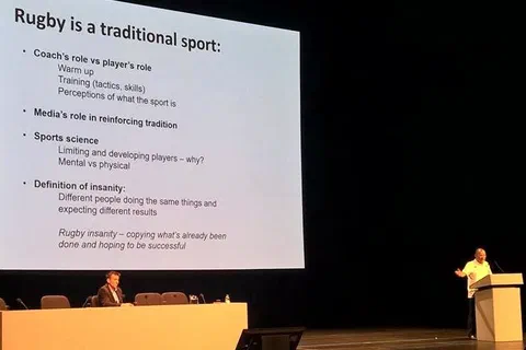 Prelegent przy mównicy w audytorium omawiający slajd o roli trenera, zawodnika i nauki o sporcie w rugby
