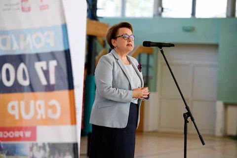 Kobieta w szarym żakiecie i okularach przemawiająca do mikrofonu na statywie podczas konferencji