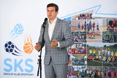 Mężczyzna w szarym garniturze przemawiający do mikrofonu na tle ścianki programu Szkolny Klub Sportowy