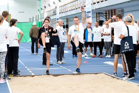 Grupa uczestników wykonujących ćwiczenia biegowe w miejscu przed skocznią w dal w hali lekkoatletycznej