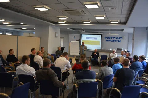 Widok sali konferencyjnej z uczestnikami słuchającymi prelegenta stojącego przy ekranie i napisie Instytut Sportu