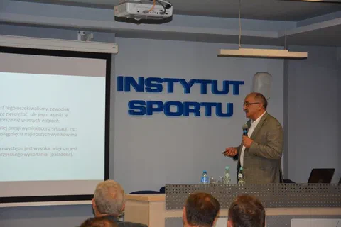Wykładowca w marynarce przemawiający do mikrofonu przed ścianą z napisem Instytut Sportu