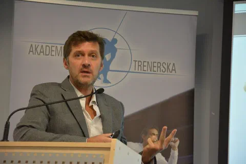 Portretowy kadr prelegenta przemawiającego przy mikrofonie na tle roll-upu Akademii Trenerskiej