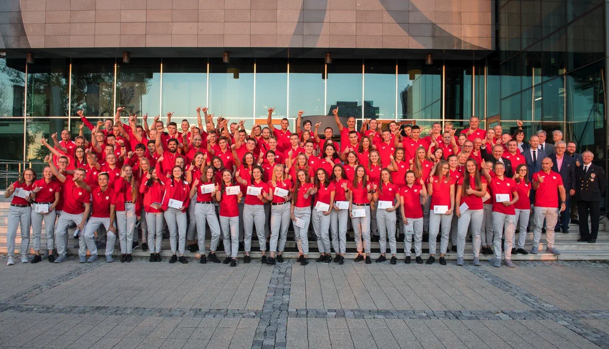 Grupowe zdjęcie Młodzieżowej Reprezentacji Polski w czerwonych koszulkach oraz trenerów i gości przed budynkiem Centrum Olimpijskiego PKOl w Warszawie.