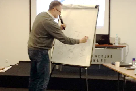 Prowadzący z mikrofonem zapisujący schemat na flipcharcie na tle ekranu projekcyjnego