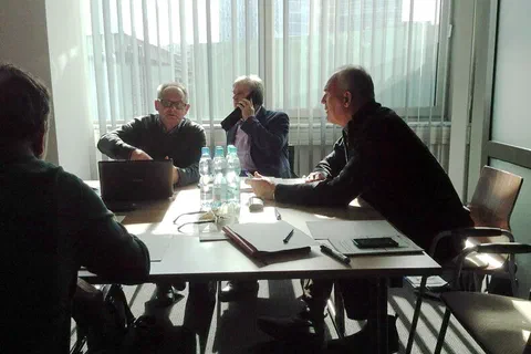 Czterech mężczyzn rozmawiających przy stole z laptopem i dokumentami na tle dużego okna
