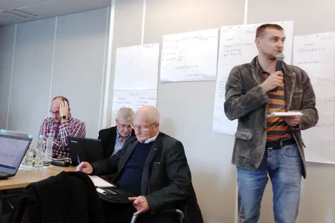 Uczestnik stojący z mikrofonem i notatkami na tle ściany z zawieszonymi arkuszami flipchartu