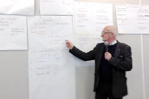 Mężczyzna z mikrofonem wskazujący długopisem wpisy na arkuszach zawieszonych na ścianie