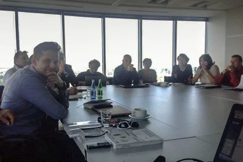 Uczestnicy siedzący wzdłuż stołu konferencyjnego na tle wysokich okien sali