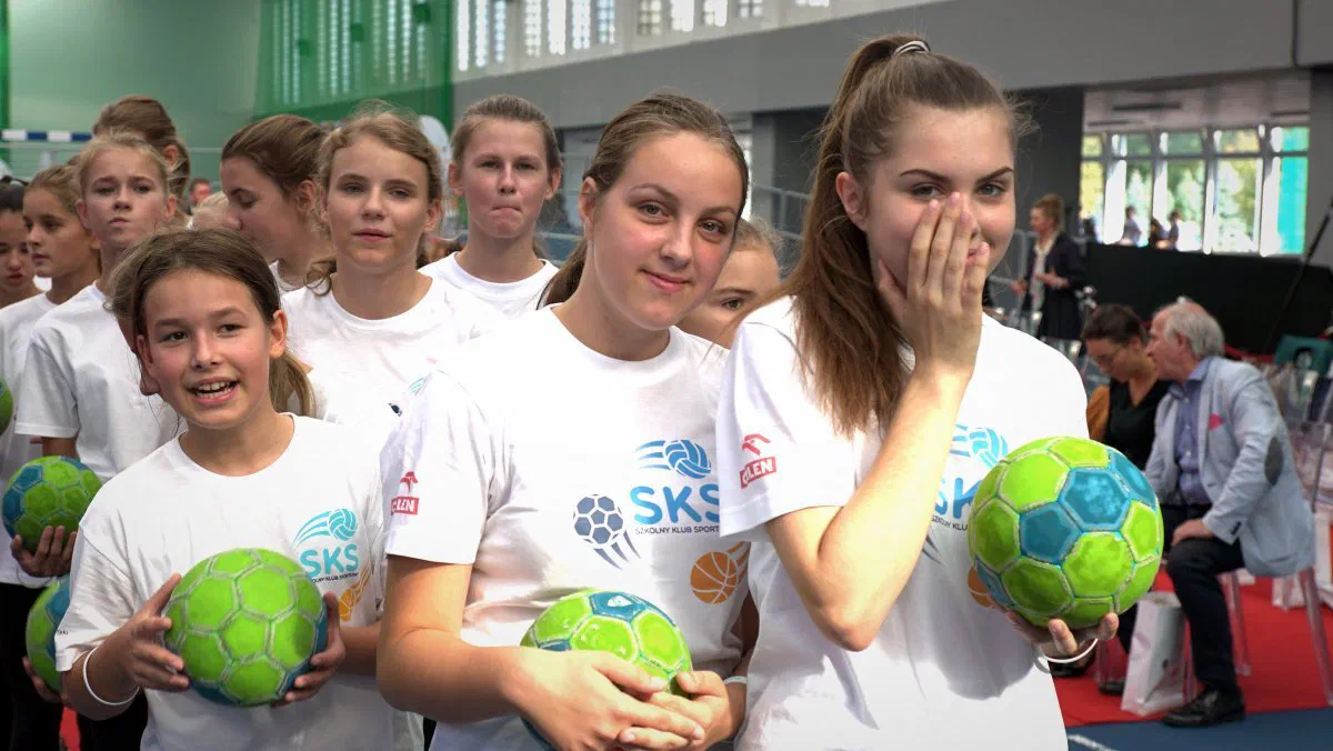 Uczennice w koszulkach programu Szkolny Klub Sportowy trzymające zielone piłki podczas zajęć w hali sportowej.