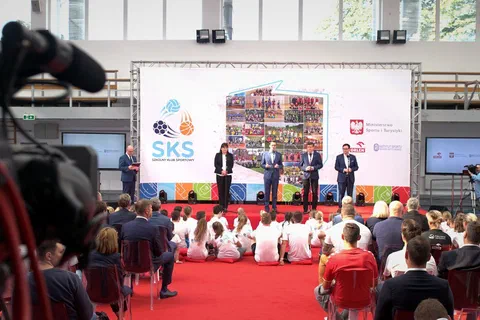 Wystąpienie na scenie przed ścianką programu SKS z dziećmi siedzącymi na podłodze hali i kamerą na pierwszym planie