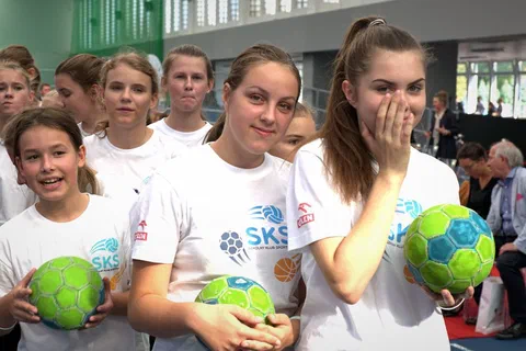 Grupa uśmiechniętych uczennic w białych koszulkach SKS trzymających zielone piłki do piłki ręcznej