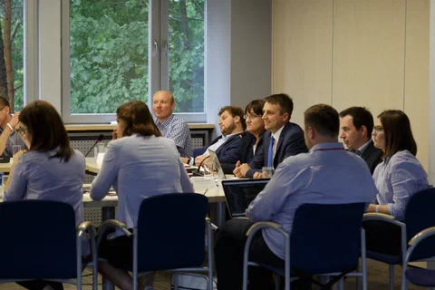 Uczestnicy konferencji siedzący naprzeciwko siebie przy długich stołach z mikrofonami w sali z oknami