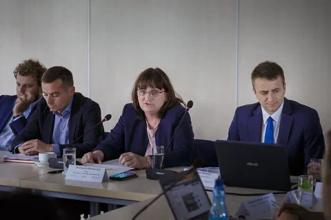 Kobieta w okularach przemawiająca do mikrofonu obok trzech mężczyzn przy stole prezydialnym