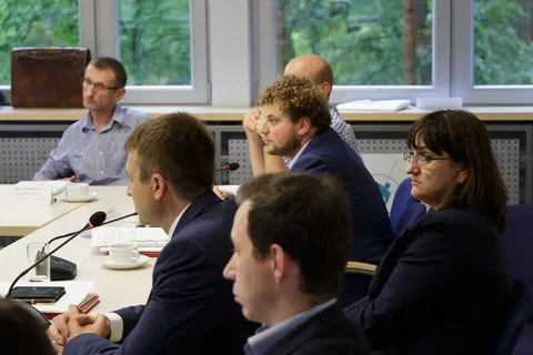 Pionowy kadr uczestników siedzących wzdłuż stołu konferencyjnego na tle zieleni za oknem