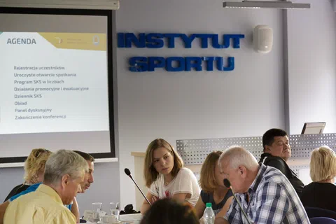 Uczestnicy siedzący przy stole pod ekranem z agendą i napisem Instytut Sportu na ścianie