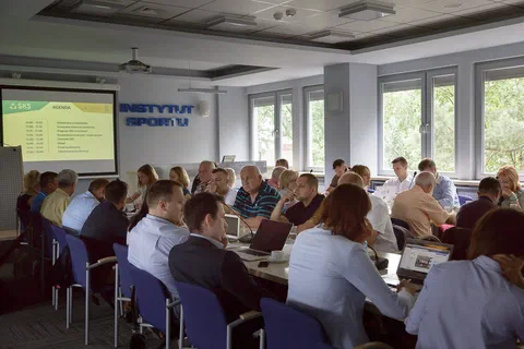 Szeroki widok sali z uczestnikami siedzącymi w podkowę i spoglądającymi na ekran z agendą