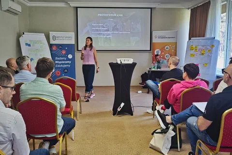 Uczestniczka prezentująca na środku sali obok ekranu z hasłem Prototypowanie i flipchartów