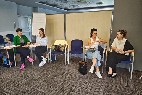 Cztery kobiety siedzące na krzesłach z pulpitami i dyskutujące w parach na tle flipchartu