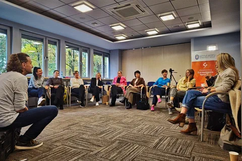 Uczestniczki siedzące w półkolu pod oknami i słuchające prowadzącego siedzącego po prawej stronie
