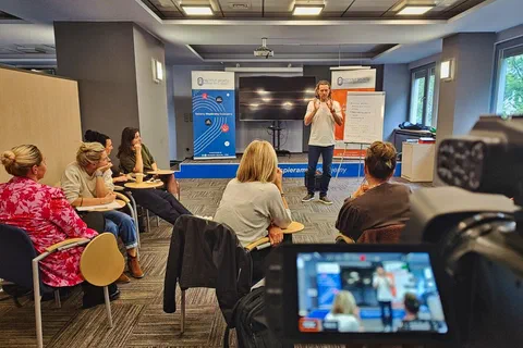 Widok zza kamery wideo na prelegenta stojącego przy flipcharcie przed grupą uczestniczek
