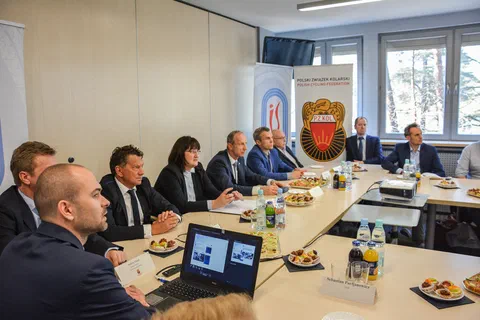 Widok sali obrad z laptopem na pierwszym planie i uczestnikami siedzącymi przy stołach