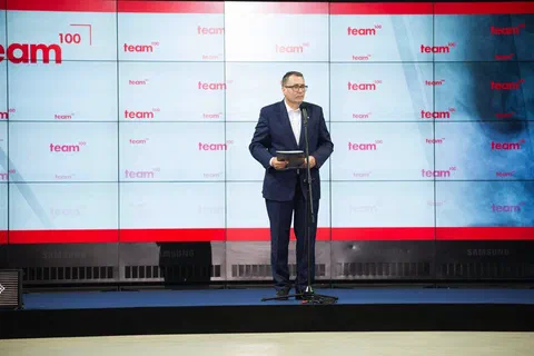 Prelegent w garniturze przemawiający przy mikrofonie na scenie na tle jasnej ścianki z logo programu Team100