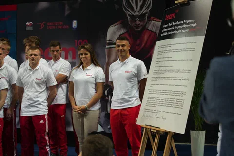 Grupa młodych sportowców w biało-czerwonych strojach stojąca obok planszy z kodeksem etyki beneficjenta programu Team100