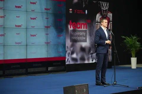 Mężczyzna w granatowym garniturze przemawiający do mikrofonu na scenie obok plakatu z hasłem Wspieramy zwycięzców