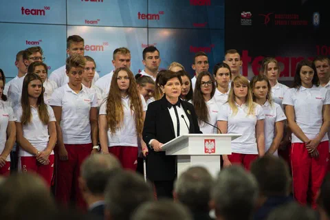 Wystąpienie przy mównicy z godłem państwowym na tle grupy młodych zawodników stojących na scenie