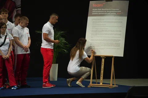 Zawodniczka klęcząca przy sztaludze i składająca podpis na planszy kodeksu etyki beneficjenta Team100