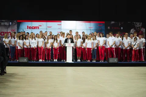 Szeroki kadr sceny z przemawiającą przy mównicy i stojącymi za nią w dwóch rzędach młodymi sportowcami